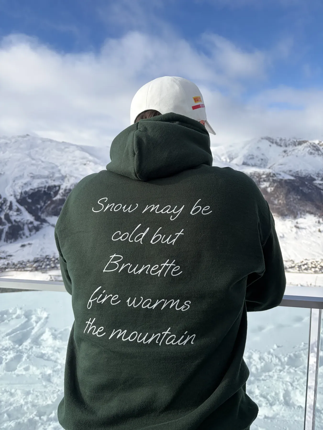 Sciatore di spalle con la felpa verde, frase bianca sul retro, davanti alle montagne innevate